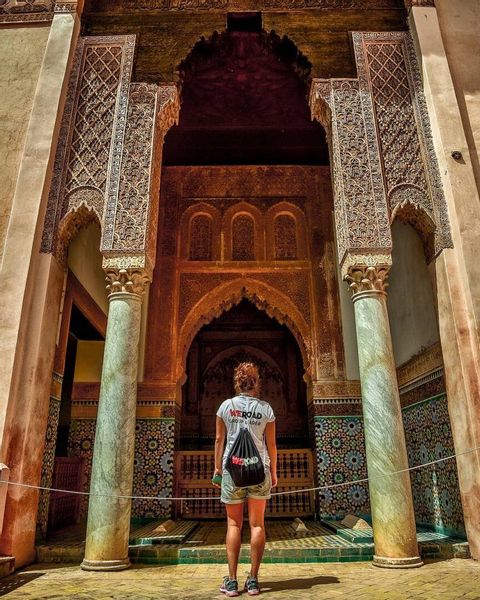 Una mujer con camiseta y mochila de WeRoad de pie mirando hacia un gran arco ornamentado con tallas detalladas y mosaicos de azulejos.