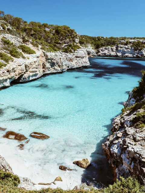 Una vista desde un acantilado alto de una cala apartada con agua turquesa transparente, una pequeña playa de arena blanca y costas rocosas.