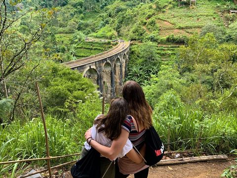 Dos mujeres de un viaje en grupo de WeRoad se abrazan mientras miran un puente de arco de piedra en un valle exuberante y verde.