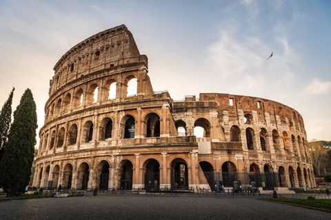 El antiguo anfiteatro Coliseo en Roma, visto desde una plaza de adoquines vacía al amanecer bajo un cielo parcialmente nublado.