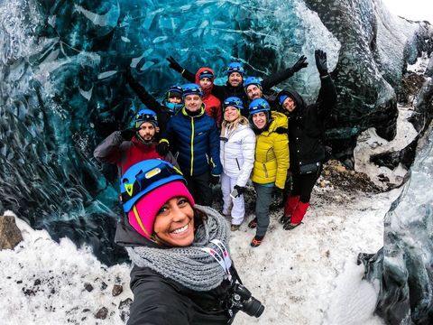 Un viaggio di gruppo WeRoad, con caschi e giacche invernali, sorride per un selfie all'interno di una grotta di ghiaccio blu traslucida.