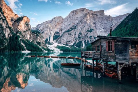 A wooden boathouse on stilts and rowboats on a calm lake, with snow-capped mountains reflected in the water.