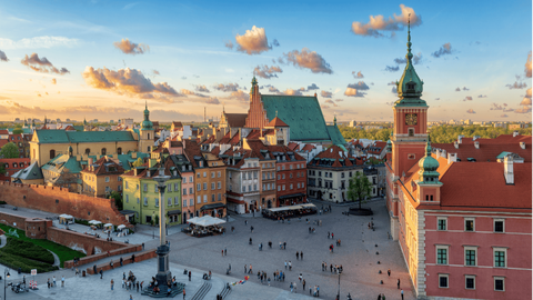 Eine Luftaufnahme eines historischen europäischen Altstadtplatzes bei Sonnenuntergang, mit Menschen, die zwischen bunten Gebäuden und einem markanten Glockenturm spazieren.