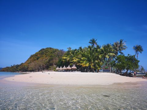 Ein Blick vom klaren, seichten Wasser einer tropischen Insel mit einem weißen Sandstrand, Palmen und einem bewaldeten Hügel unter blauem Himmel.