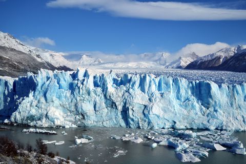 Un'imponente parete di ghiacciaio blu si affaccia su uno specchio d'acqua con blocchi di ghiaccio galleggianti, sullo sfondo montagne innevate.