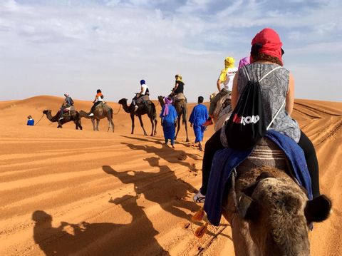 Un viaje en grupo de WeRoad monta en camellos en una caravana a través de dunas de arena naranjas bajo un cielo parcialmente nublado.