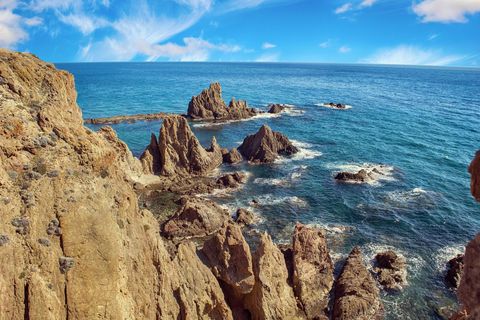 Formaciones rocosas dentadas sobresalen del océano turquesa, vistas desde un acantilado bajo un cielo azul parcialmente nublado.