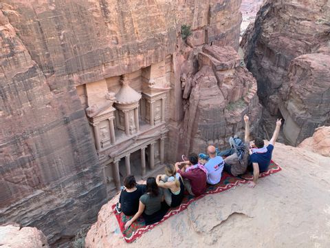 Un grupo de WeRoad, sentado sobre una alfombra en la cima de un acantilado, contempla un edificio antiguo tallado en la roca roja.