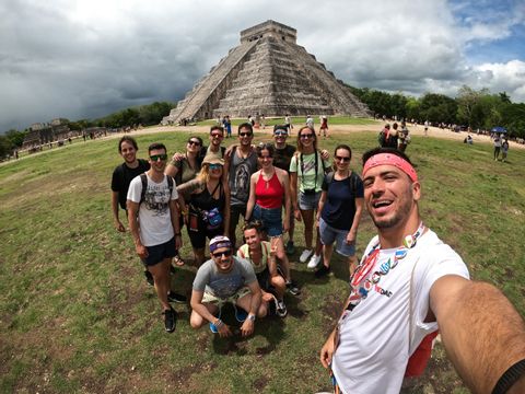 Un viaje en grupo de WeRoad se toma una selfie sonriente en un campo de hierba frente a una gran pirámide de piedra antigua bajo un cielo nublado.