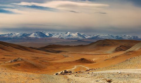 Una caravana de personas y ganado se mueve por un valle árido por un camino de tierra, con montañas nevadas al fondo.