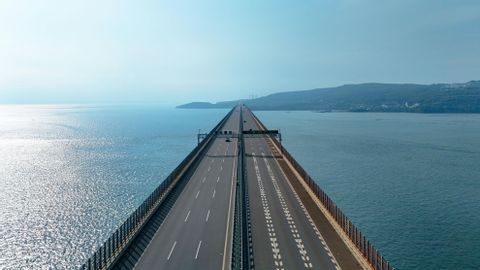 A long highway bridge extends over the sea towards a distant, hazy shoreline.
