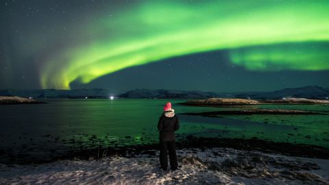 Una persona con un gorro rosa de pie en una orilla nevada observando la aurora boreal verde iluminar el cielo nocturno sobre una gran masa de agua.