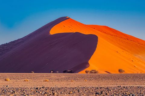 A large orange sand dune casts a deep shadow across itself in a rocky desert landscape under a clear blue sky.