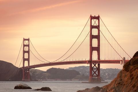 Eine große rote Hängebrücke überspannt ein Gewässer mit Hügeln im Hintergrund unter einem warm getönten Himmel bei Sonnenuntergang.
