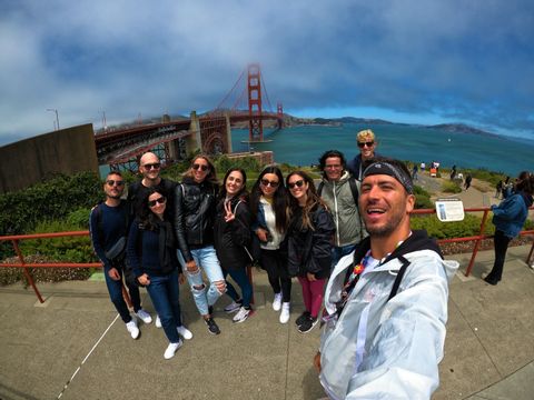 Un gruppo WeRoad si fa un selfie da un punto panoramico, con un grande ponte sospeso rosso e il mare sullo sfondo.