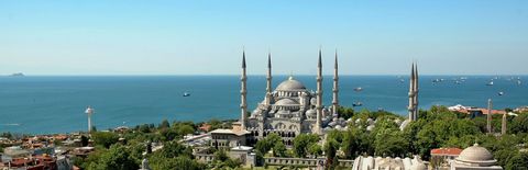 Una grande moschea con molte cupole e sei minareti che si affaccia su un mare pieno di navi sotto un cielo azzurro e limpido.