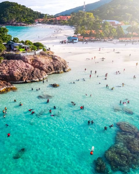 An aerial view of people swimming and snorkeling in clear turquoise water next to a white sand beach backed by lush green hills.
