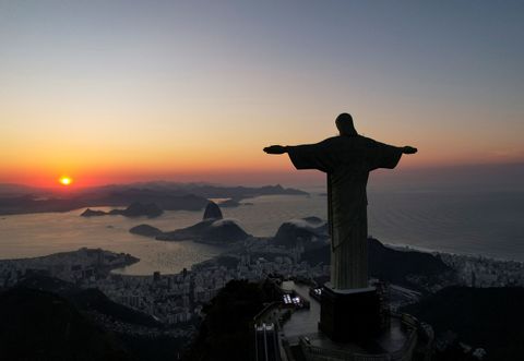 The silhouette of a large statue with outstretched arms overlooks a coastal city, a bay, and mountains during a colorful sunrise.