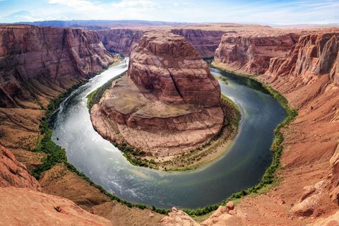 Un fiume scorre a forma di U attorno a una grande formazione rocciosa all'interno di un vasto canyon rosso sotto un cielo parzialmente nuvoloso.