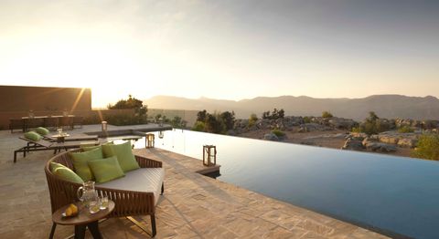 Un patio moderno con una piscina infinita y mobiliario de salón con vistas a un paisaje montañoso rocoso al atardecer.