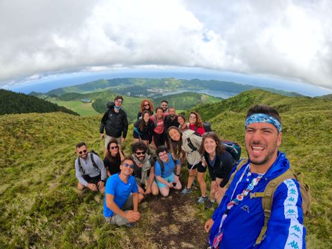 Un grupo de viaje de WeRoad se toma una selfie en una colina cubierta de hierba con vistas a un lago de cráter y al océano bajo un cielo nublado.