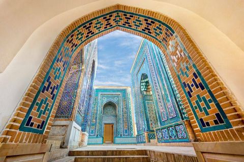 Ein Blick durch einen großen Ziegel- und Fliesenbogen auf einen Innenhof mit Gebäuden, die mit aufwendigen blauen und türkisen Mosaiken bedeckt sind.