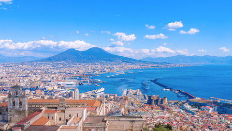 Elevated panoramic view of a coastal city harbor and bay with a large twin-peaked mountain under a blue sky.