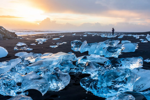 De gros morceaux de glace cristalline sont éparpillés sur une plage de sable noir, avec une personne observant l'océan au coucher du soleil.