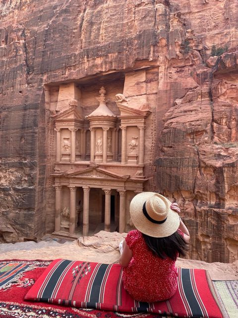 Una mujer con vestido rojo y sombrero de paja se sienta sobre una alfombra, observando un gran templo tallado en un acantilado de roca roja.