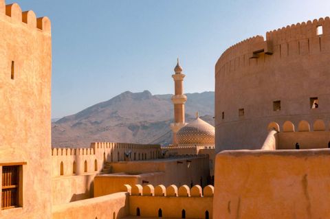 A mosque's golden dome and minaret rise above the sand-colored walls of a fortress, with distant mountains under a clear sky.
