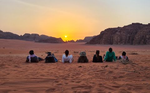Un grupo de WeRoad en un viaje contempla el atardecer sobre la arena roja del desierto, con formaciones rocosas distantes al fondo.