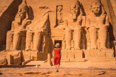 A woman in a red dress and straw hat walks towards a large ancient temple carved into a cliff with colossal seated statues.