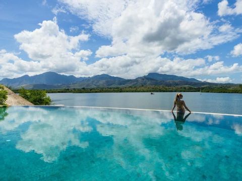 Eine Frau am Rande eines Infinity-Pools blickt über eine ruhige Bucht auf ferne Berge unter einem teilweise bewölkten Himmel.