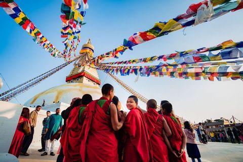 A group of people in red robes look towards a large white stupa with colorful prayer flags strung from its spire against a blue sky.