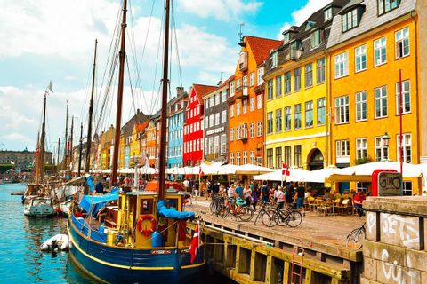 Edificios coloridos bordean un canal donde hay veleros atracados, con gente caminando y sentada en cafeterías a lo largo del muelle frente al mar.