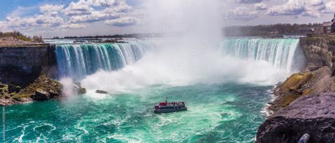 Un barco turístico navega sobre turbulentas aguas turquesas cerca de la base de una enorme cascada en forma de herradura, bajo un cielo nublado.