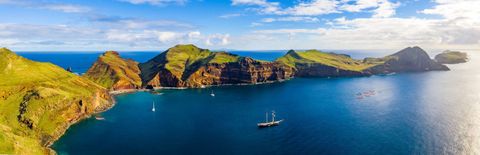 A panoramic aerial view of a rugged, green coastline and deep blue sea, with a large sailing ship and other boats.