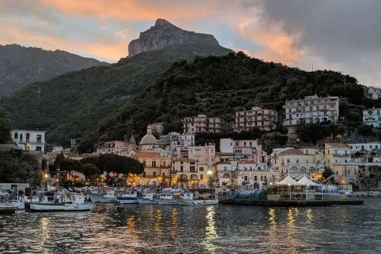 Un pueblo costero con su puerto lleno de barcos, iluminado al anochecer con una gran montaña arbolada al fondo.