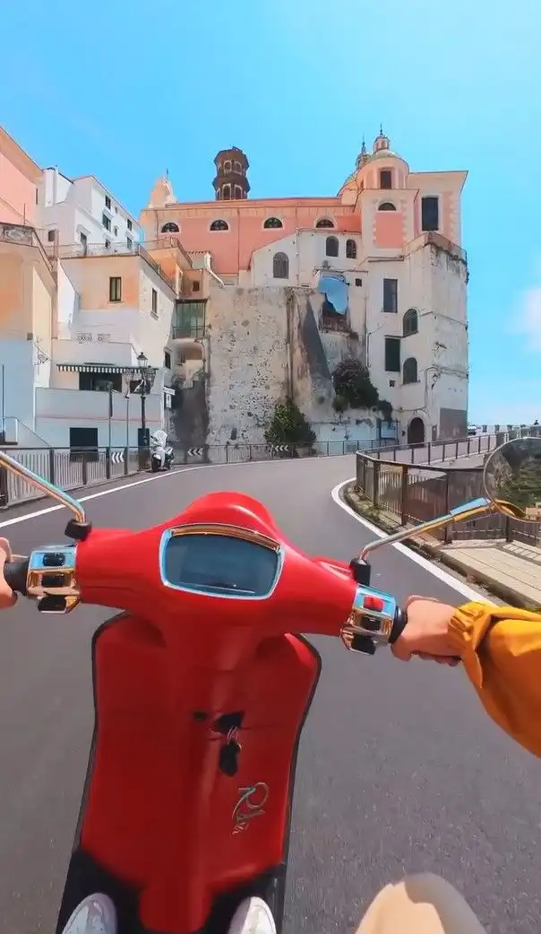 Paseo en primera persona en scooter roja por una carretera sinuosa entre edificios históricos en un pueblo costero soleado.