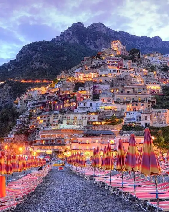 Pueblo iluminado en la ladera al atardecer, visto desde una playa de guijarros con sombrillas plegadas y tumbonas.