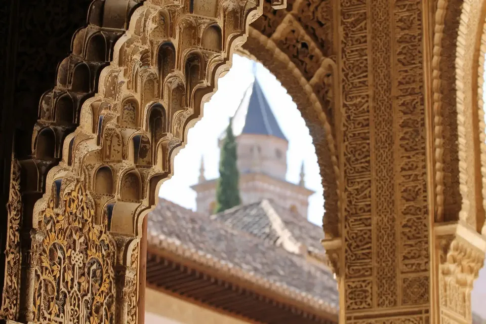Primer plano de un arco ornamentado con tallas detalladas, enmarcando una vista de una torre y un tejado de tejas al fondo.