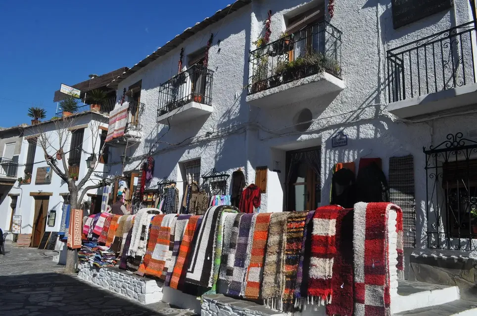 Alfombras tejidas coloridas y ropa cuelgan en exhibición en una calle frente a un edificio blanco bajo un cielo azul claro.