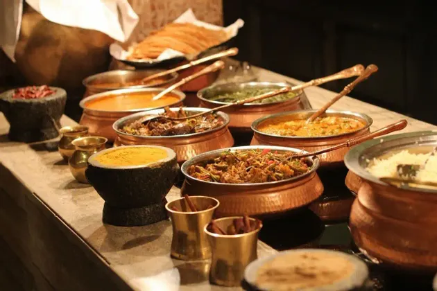 Un buffet di varie pietanze indiane, inclusi curry e riso, servite in tradizionali ciotole di rame su un piano di lavoro in pietra.