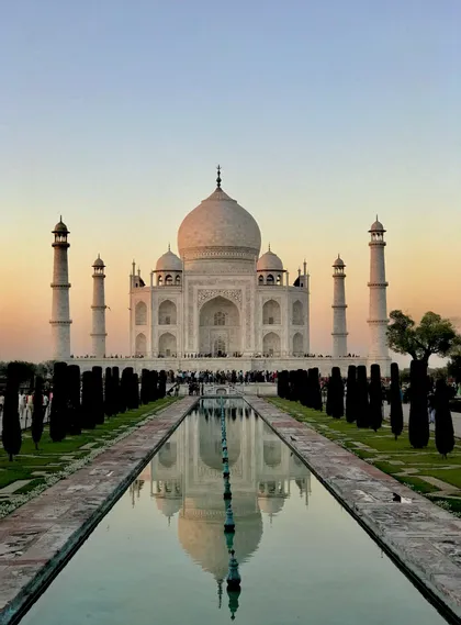 Una vista simmetrica del Taj Mahal che si riflette in una lunga vasca al tramonto, con giardini verde scuro che costeggiano l'acqua.