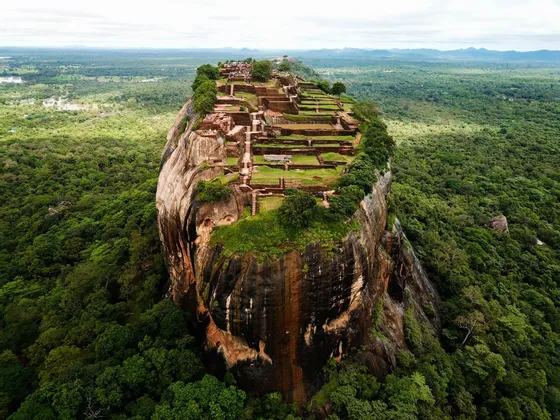 Vista aerea di antiche rovine e giardini a terrazze in cima a una massiccia fortezza rocciosa che emerge da una fitta giungla verde.