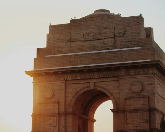 La cima del monumento in pietra India Gate illuminata dalla calda luce del sole al tramonto contro un cielo velato.