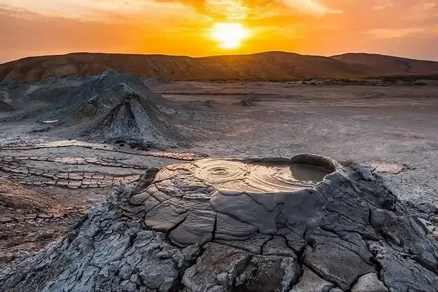 Vulcani di fango ribollenti su un campo di terra screpolata con una pozza in primo piano sotto un tramonto dorato.