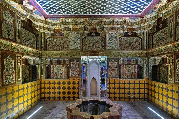 L'interno di una stanza sontuosa con pareti e soffitto ricoperti da intricati mosaici floreali e geometrici sopra piastrelle gialle, e una fontana centrale in mattoni.