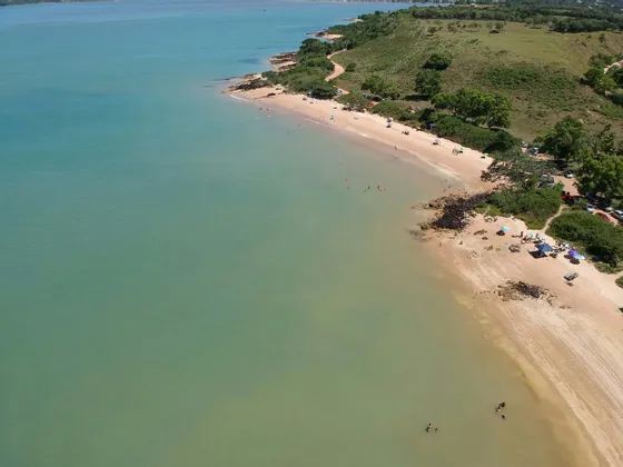 Vista aerea di una spiaggia sabbiosa lungo una costa verde e collinare, con persone che nuotano in acque calme e turchesi.