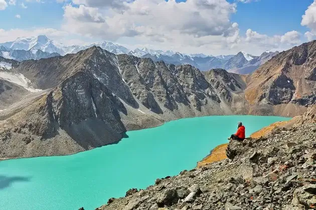 Eine Person in einer roten Jacke sitzt auf einer felsigen Klippe mit Blick auf einen helltürkisen Alpensee, umgeben von Bergen unter einem bewölkten Himmel.